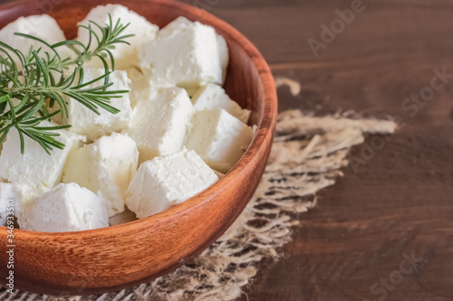 slices of feta cheese in a wooden bowl and sprigs of rosemary close-up. background with diced feta cheese and sprigs of fresh rosemary. feta cheese close-up and fresh greens.