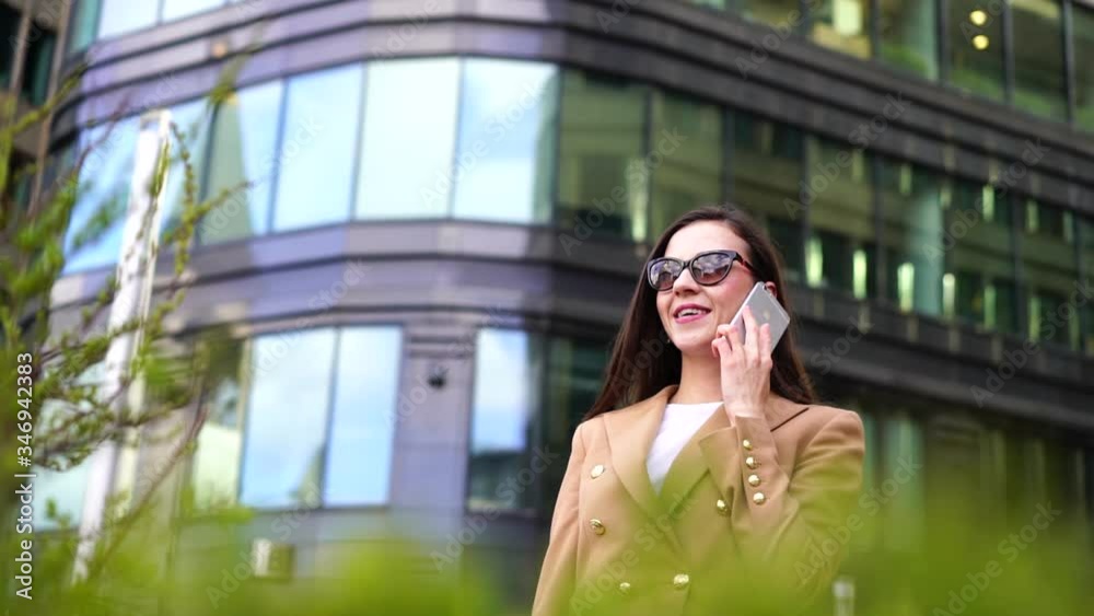 Close-up portrait of a woman, walking around the city against the background of a European office building, wearing a stylish beige jacket and dark glasses. She's on the phone and laughing. Close-up portrait of a woman, walking around the city against the background of a European office building, wearing a stylish beige jacket and dark glasses. She's on the phone and laughing.