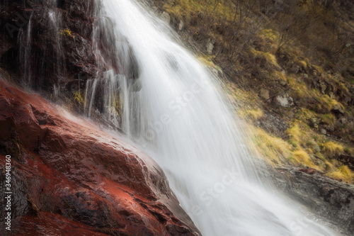 Stunning close up details of powerful waterfall cascading down the red  wet rocks and soft  background sunlit golden grass