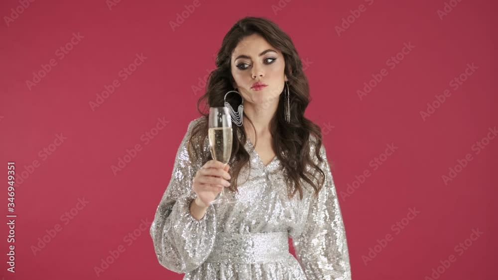 A sad young woman in bright sequins dress is holding a glass of champagne isolated over pink wall background A sad young woman in bright sequins dress is holding a glass of champagne isolated over pink wall background