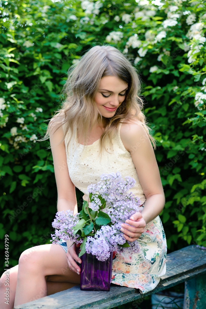 A young woman sits on a bench in a summer garden with a bouquet of lilacs A young woman sits on a bench in a summer garden with a bouquet of lilacs