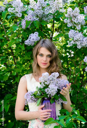 Beautiful girl with green eyes in a summer dress with a bouquet of lilacs in a blooming garden
