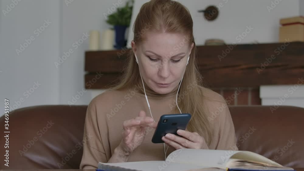 Concentrated Caucasian woman in earphones scrolling smartphone screen. Portrait of serious adult businesswoman checking information online. Wireless technologies, lifestyle, business. Concentrated Caucasian woman in earphones scrolling smartphone screen. Portrait of serious adult businesswoman checking information online. Wireless technologies, lifestyle, business.