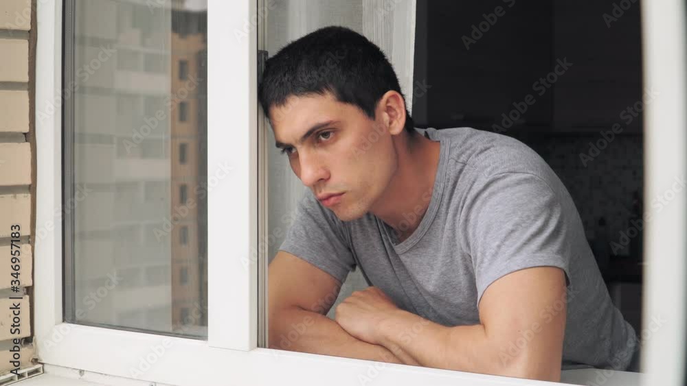 A sad looking guy standing at the window. Depression and problems during the period of isolation and quarantine A sad looking guy standing at the window. Depression and problems during the period of isolation and quarantine