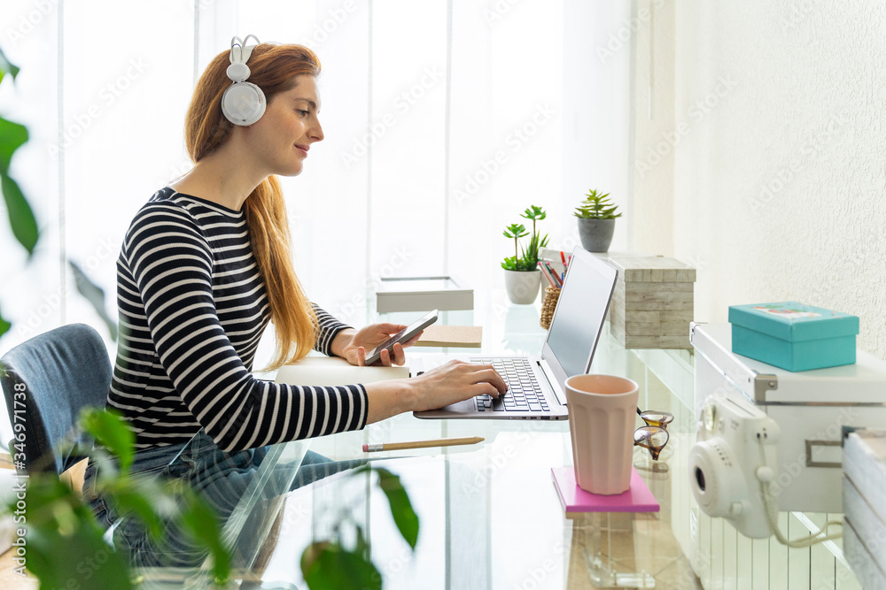 Young woman wearing headphones using laptop and smartphone at desk at home Young woman wearing headphones using laptop and smartphone at desk at home