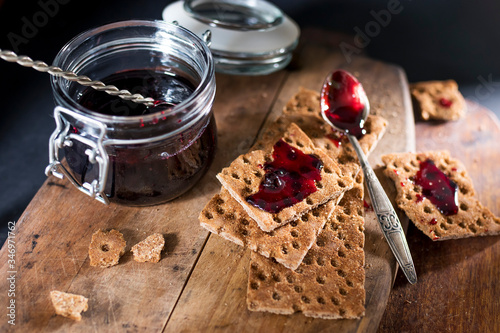 Open jar with blackcurrant jam on a wooden cutting board. Dry biscuits spread with jam, crumbs and a spoon in jam. Place for text.