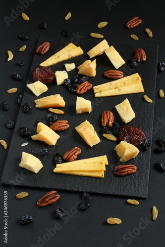 Pieces of parmesan cheese and gouda cheese with fenugreek, blue and gold raisins and dried apricots on black stone plate on black table. Closeup