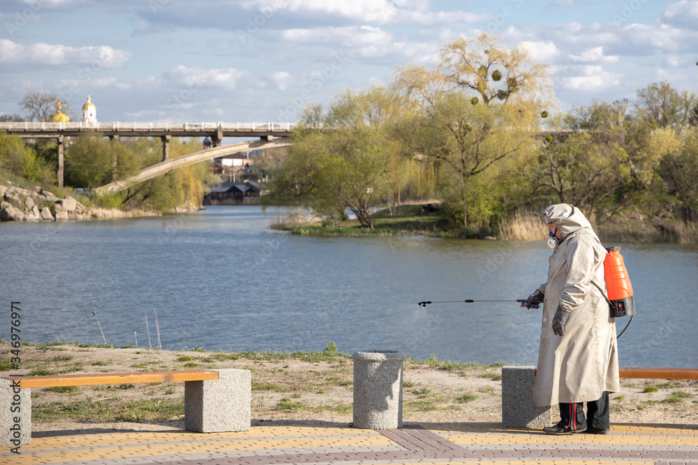 Fototapeta premium Bila Tserkva, Ukraine - April 20, 2020: A man in a gray coat treats the area with a cleaning solution. Shops, trash cans processing from Covid-19. The Covid-19 Epidemic. Pandemic. Beach. River.