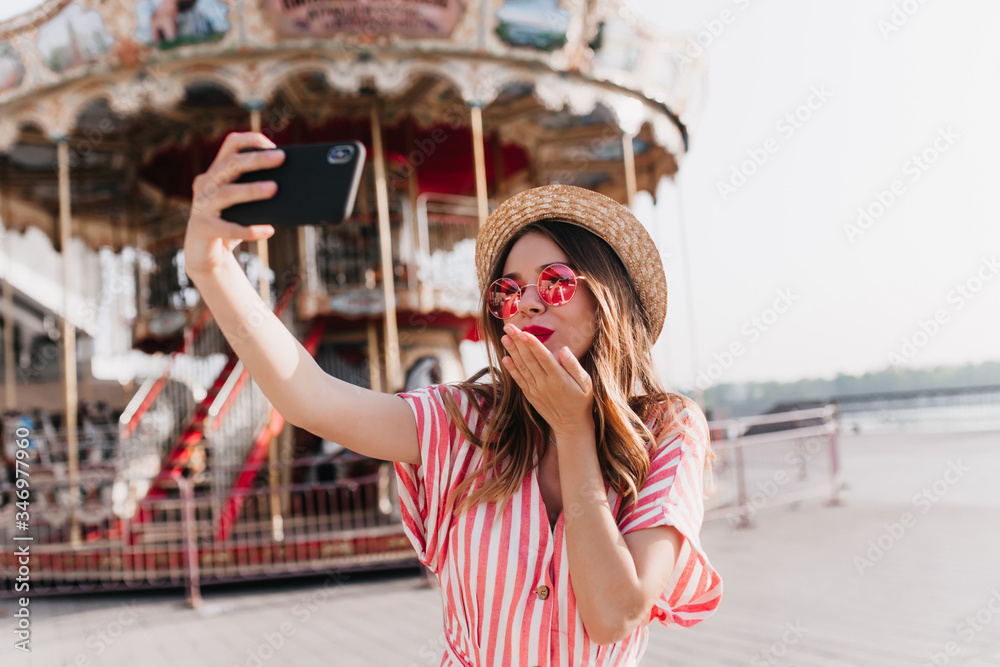 Inspired caucasian girl in straw hat sending air kiss for selfie. Outdoor shot of fascinating woman in pink sunglasses taking picture of herself near carousel.
