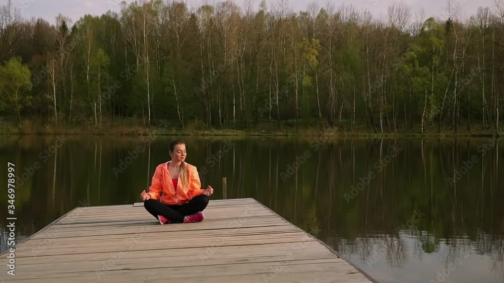 Girl is practicing yoga technique. She is sitting on the pier in lotus pose by the lake Girl is practicing yoga technique. She is sitting on the pier in lotus pose by the lake