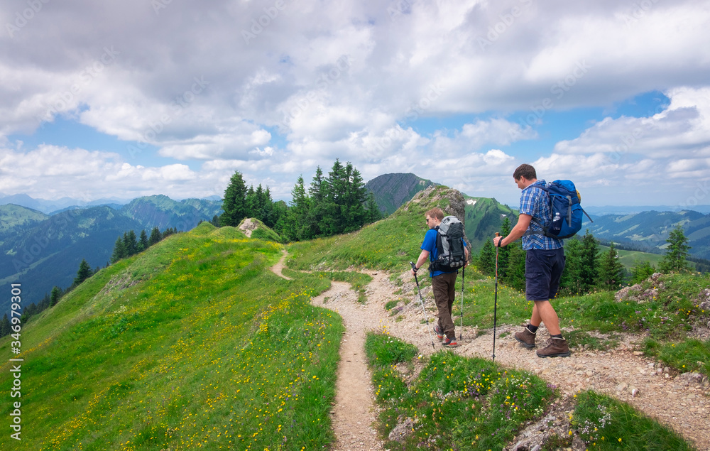 Two men hiking along footpath with flowers and summer meadow in the mountain of Nagelfluhkette, clouds in the sky, Allgäu Oberstaufen Germany.