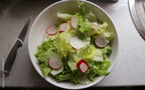 Freshly washed green salad with sliced red radish in white, ceramic bowl on the kitchen counter
