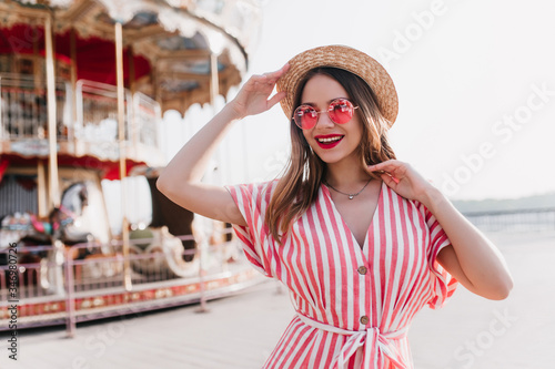 Blithesome blonde girl in striped dress posing with pleasure in amusement park. Outdoor shot of smiling fashionable lady in summer hat standing beside carousel.
