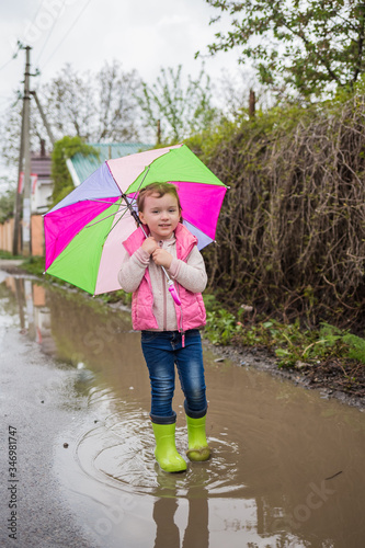 A little girl with a colorful umbrella and green boots is standing in a puddle.