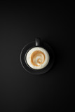 latte art shape on milk foam in black cup and saucer on a black surface in the center of the frame. Flat lay.