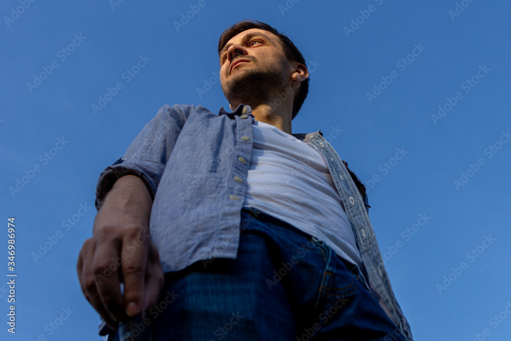 a young man wearing jeans, a white T-shirt and a blue shirt stands in a field against a blue cloudless sky, bottom view. a young man wearing jeans, a white T-shirt and a blue shirt stands in a field against a blue cloudless sky, bottom view.
