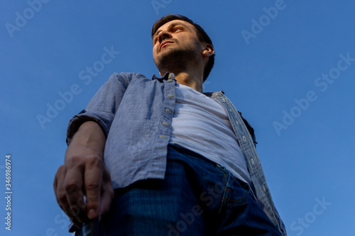 a young man wearing jeans, a white T-shirt and a blue shirt stands in a field against a blue cloudless sky, bottom view.