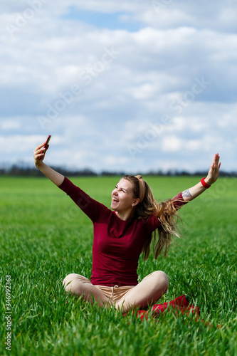 Young beautiful woman student with a phone in her hands sitting on the grass. Girl takes selfies and takes selfie pictures. She smiles and enjoys a warm day. Concept photo on smartphone