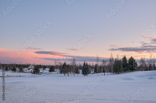 Landscape. The hills are covered with snow with growing trees, a small closed gazebo and a sky with clouds painted with the colors of the sunset.