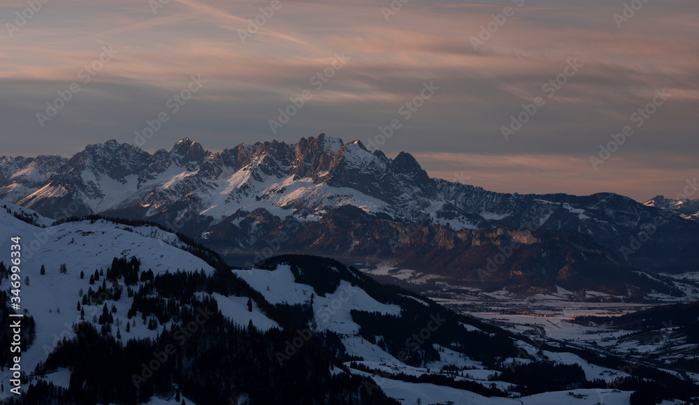 Mountains of mountain range Wilder Kaiser at Fieberbrunn during sunset in winter with snow, forest and valley, clouds in the sky, Tyrol Austria.