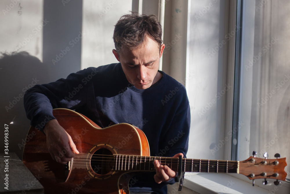 Obraz premium Guy with a guitar in the kitchen. Natural light comes to the musician from the window. Lesson playing the guitar. Unknown talented musician. Playing an acoustic guitar. Brutal man shows skill.