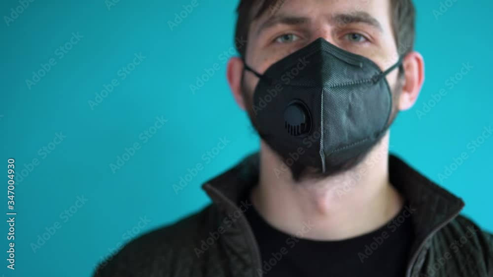 Portrait of bearded Caucasian brunet man with freckles. Attractive male with short brown hair in a black jacket puts on and takes off a protective face mask against blue wall background close up. Portrait of bearded Caucasian brunet man with freckles. Attractive male with short brown hair in a black jacket puts on and takes off a protective face mask against blue wall background close up.
