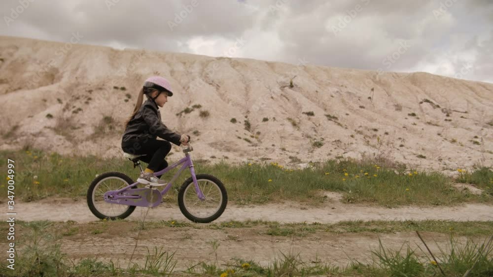 Cute little girl in helmet ride a bicycle on country road between white hill and forest. Steadycam slow motion shot Cute little girl in helmet ride a bicycle on country road between white hill and forest. Steadycam slow motion shot