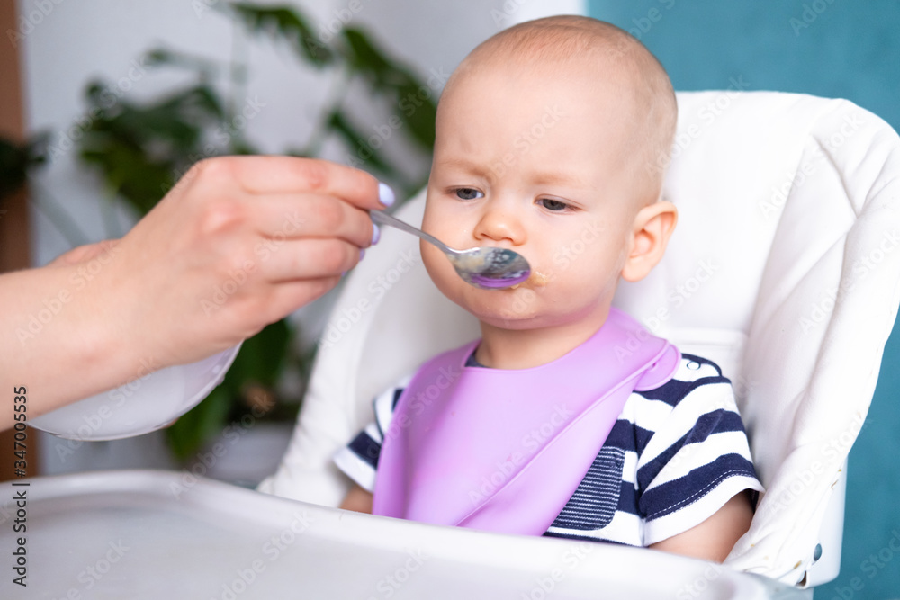 Sad baby eating tasteless food. Mom feeds little caucasian child on highchair. healthy porridge, mother hands with spoon. Newborn infant kids. Sad baby eating tasteless food. Mom feeds little caucasian child on highchair. healthy porridge, mother hands with spoon. Newborn infant kids.
