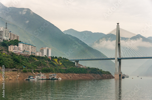 Xinling, China - May 6, 2010: Xiling gorge on Yangtze River. Part of Badong bridge with tall city buildings on slope above green water with boats. Descending cloudscape over green mountain range. photo