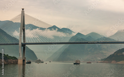 Xinling, China - May 6, 2010: Xiling gorge on Yangtze River. Part of Badong suspension bridge above green water with boats. Descending cloudscape over green mountain range landscape. photo