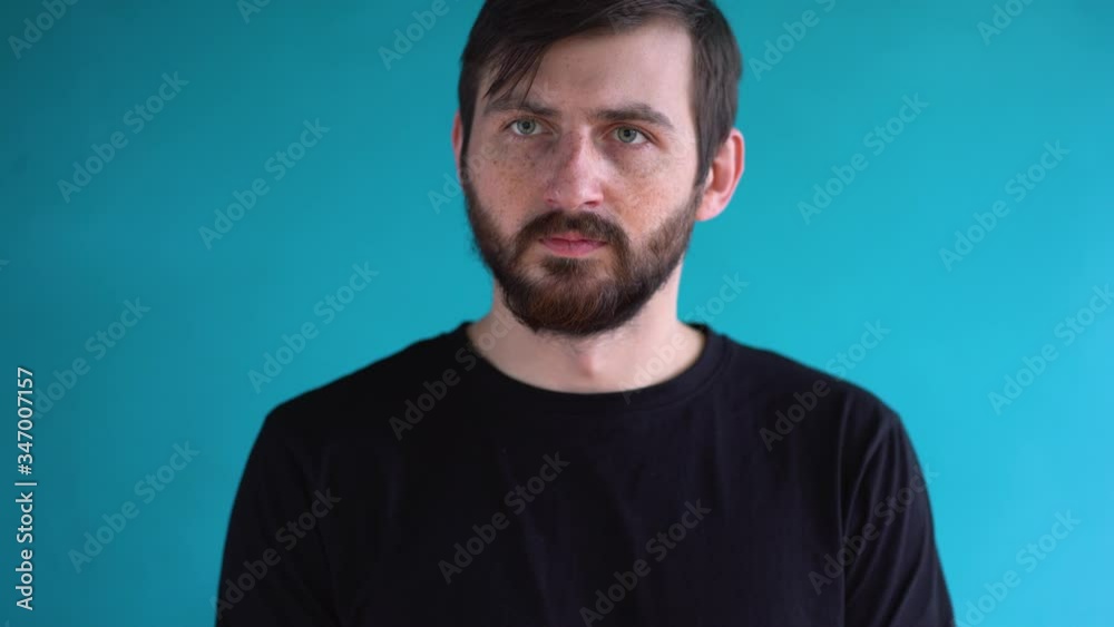 Young gorgeous man with short brown hair with freckles in black t-shirt stands in front of the camera, looks down and to the side, nods, agrees with someone against blue wall background close up. Young gorgeous man with short brown hair with freckles in black t-shirt stands in front of the camera, looks down and to the side, nods, agrees with someone against blue wall background close up.