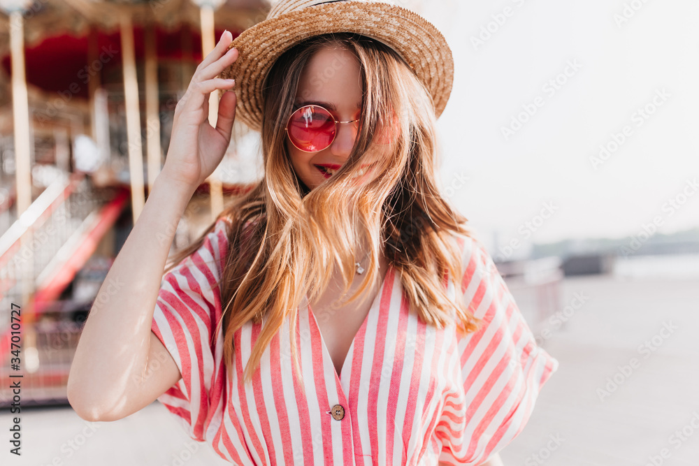 Long-haired wonderful lady wears straw hat expressing positive emotions during rest in amusement park. Outdoor photo of smiling ecstatic girl in pink sunglasses posing on city background in summer day Long-haired wonderful lady wears straw hat expressing positive emotions during rest in amusement park. Outdoor photo of smiling ecstatic girl in pink sunglasses posing on city background in summer day