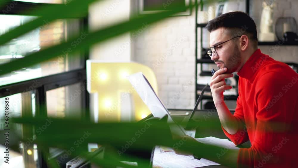 Pensive young man in eyeglasses reading data on paper documents and checking report sitting at table and working at home interior in modern apartment.Bearded male designer viewing sketches 
