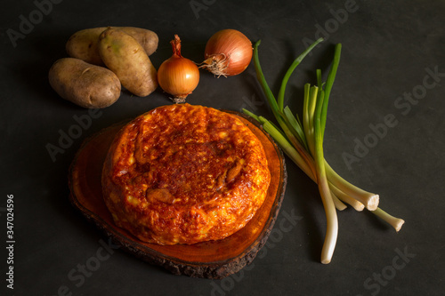 Potato omelette on a wooden board next to potatoes and onions on a black background