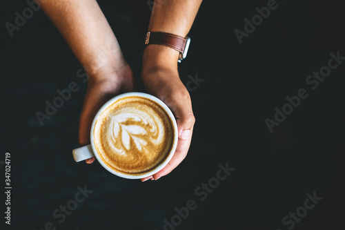 A white cup of coffee with latte art held by a young woman over a black background