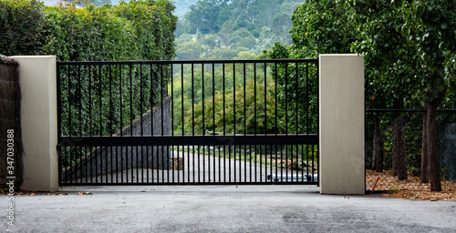 Black metal wrought iron driveway property entrance gates set in brick fence, concrete path, garden trees
