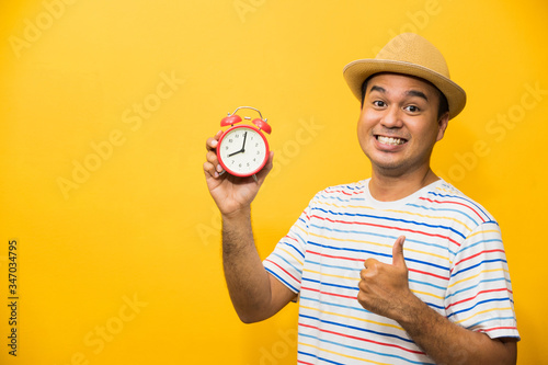 Young asian man holding the alarm clock on yellow isolated background.