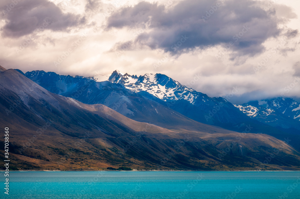 Dramatic view of Lake Pukaki with clouds gathering over the snow-capped mountain peaks. The alpine lake is famous for the amazing turquoise hues of the water and in New Zealand, South Island.