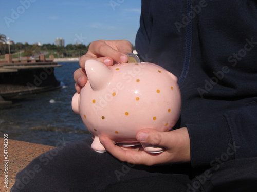 Money saving in piggy bank. A young boy puts a coin in a pink ceramic piglet piggyback on the waterfront with the city and the sea in the background lit by sunlight photo