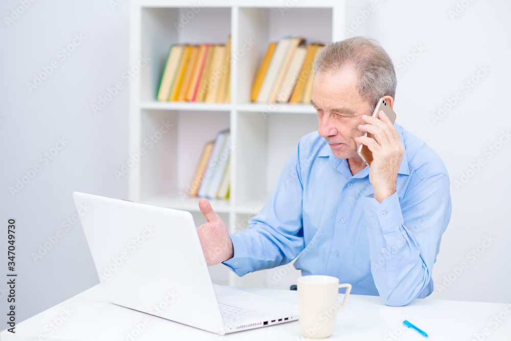Senior talking on a cell phone near a laptop on the background of shelving with books. Hardware problems, work difficulties Senior talking on a cell phone near a laptop on the background of shelving with books. Hardware problems, work difficulties