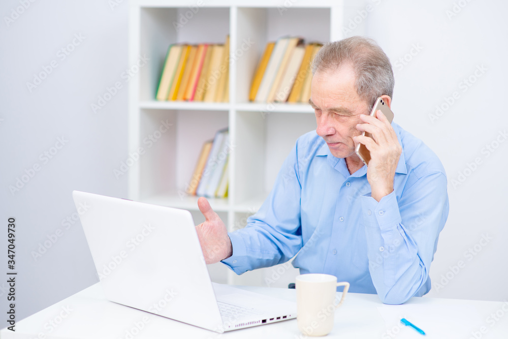 Senior talking on a cell phone near a laptop on the background of shelving with books. Hardware problems, work difficulties