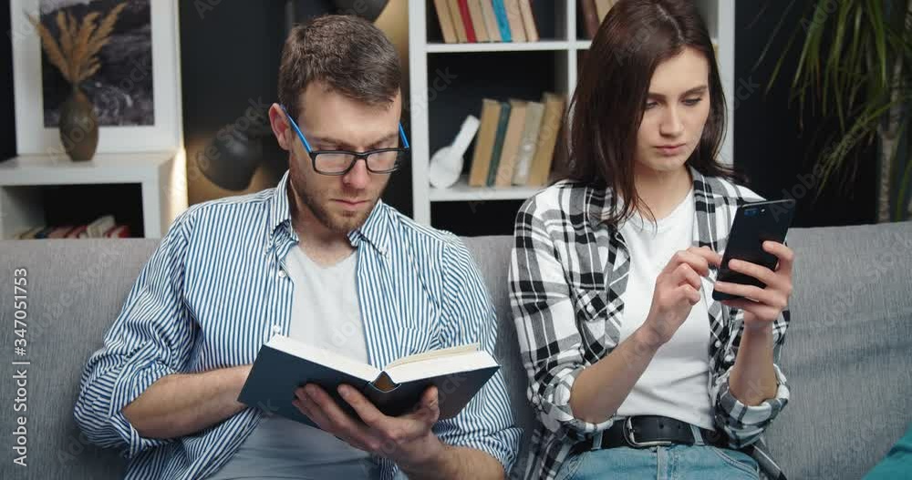 Handsome man reading interesting book while his beautiful girlfriend sitting on couch and using smartphone. Happy couple resting together at cozy apartment Handsome man reading interesting book while his beautiful girlfriend sitting on couch and using smartphone. Happy couple resting together at cozy apartment