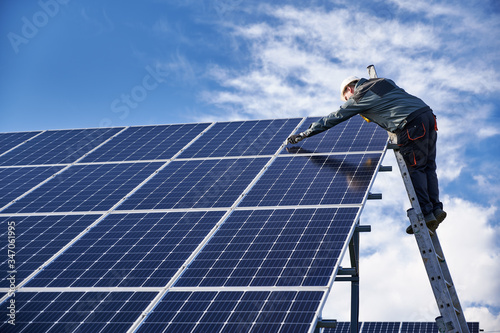 Male worker in white safety helmet standing on ladder and repairing photovoltaic solar panel station under beautiful blue cloudy sky. Concept of alternative sources of energy. photo