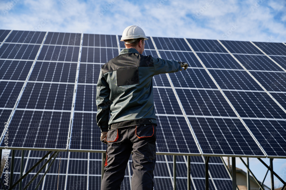 Low angle view on technician in dark blue uniform standing with his back to the camera, pointing at solar plant. Rear view. Alternative ecological source of energy concept Low angle view on technician in dark blue uniform standing with his back to the camera, pointing at solar plant. Rear view. Alternative ecological source of energy concept