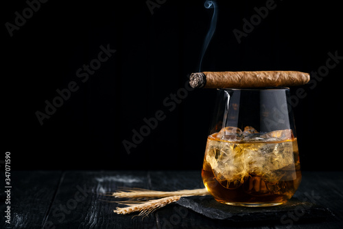 A glass of whiskey or cognac with ice cubes and a Smoking cigar lying on top, black wooden background