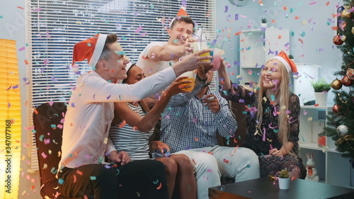 Close up of cheerful company celebrating Christmas in confetti blowing while making cheers and blowing party whistles at home.