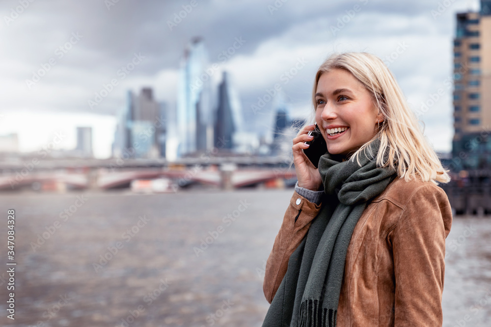 Portrait einer attraktiven, blonden Frau beim Telefonieren mit ihrem Handy vor der modernen Skyline von London