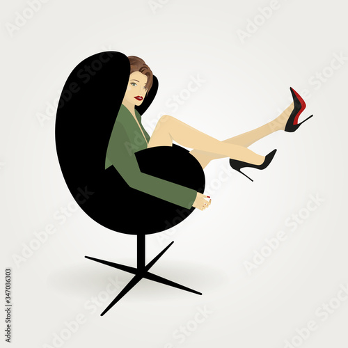 A beautiful girl with long legs in high-heeled shoes sits in a green jacket on a black armchair on a light background.