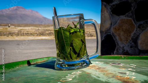 closeup of a glass mug with tea mate de coca, coca leaves and herbs infused with hot water, peruvian drink for altitude sickness, on a green barrel in front of mountains and stone wall