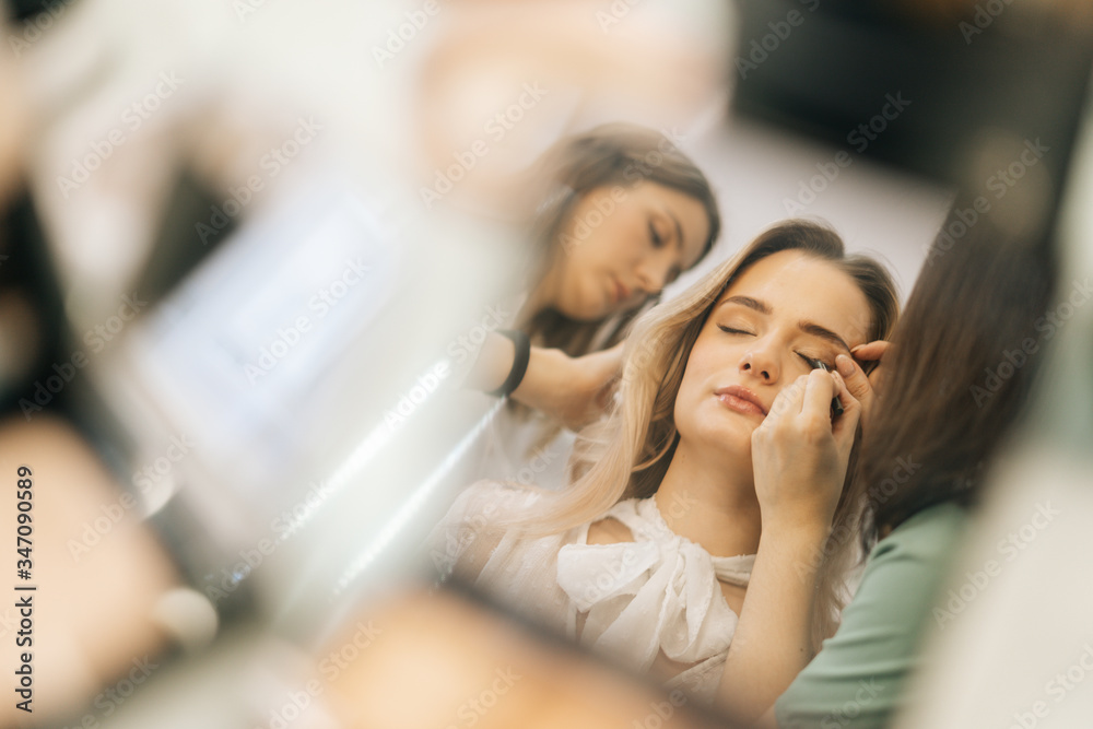 Close-up of face of young woman reflected in mirror of powder on woman table. Professional stylist and makeup artist doing makeup and style hair for young woman. Concept of backstage work. Close-up of face of young woman reflected in mirror of powder on woman table. Professional stylist and makeup artist doing makeup and style hair for young woman. Concept of backstage work.